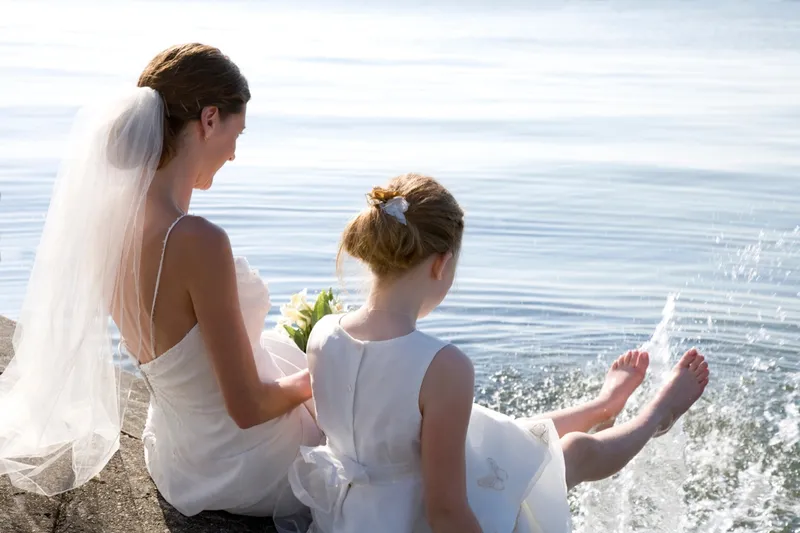 A bride and a flower girl in a white dress splashing their feet in the water at a beach wedding