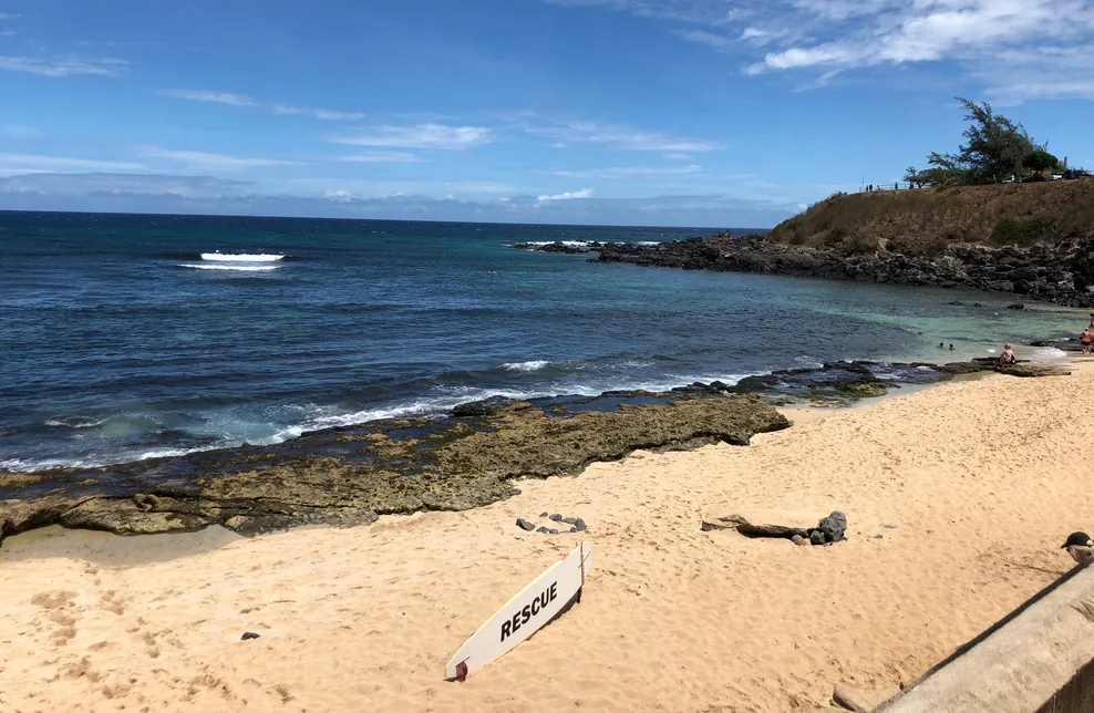 Rocky Hawaiian cove with a lifeguard rescue board on the sand and surfers on the break beyond