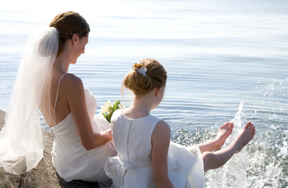 Bride and young flower girl sitting by the water, the child splashing her feet